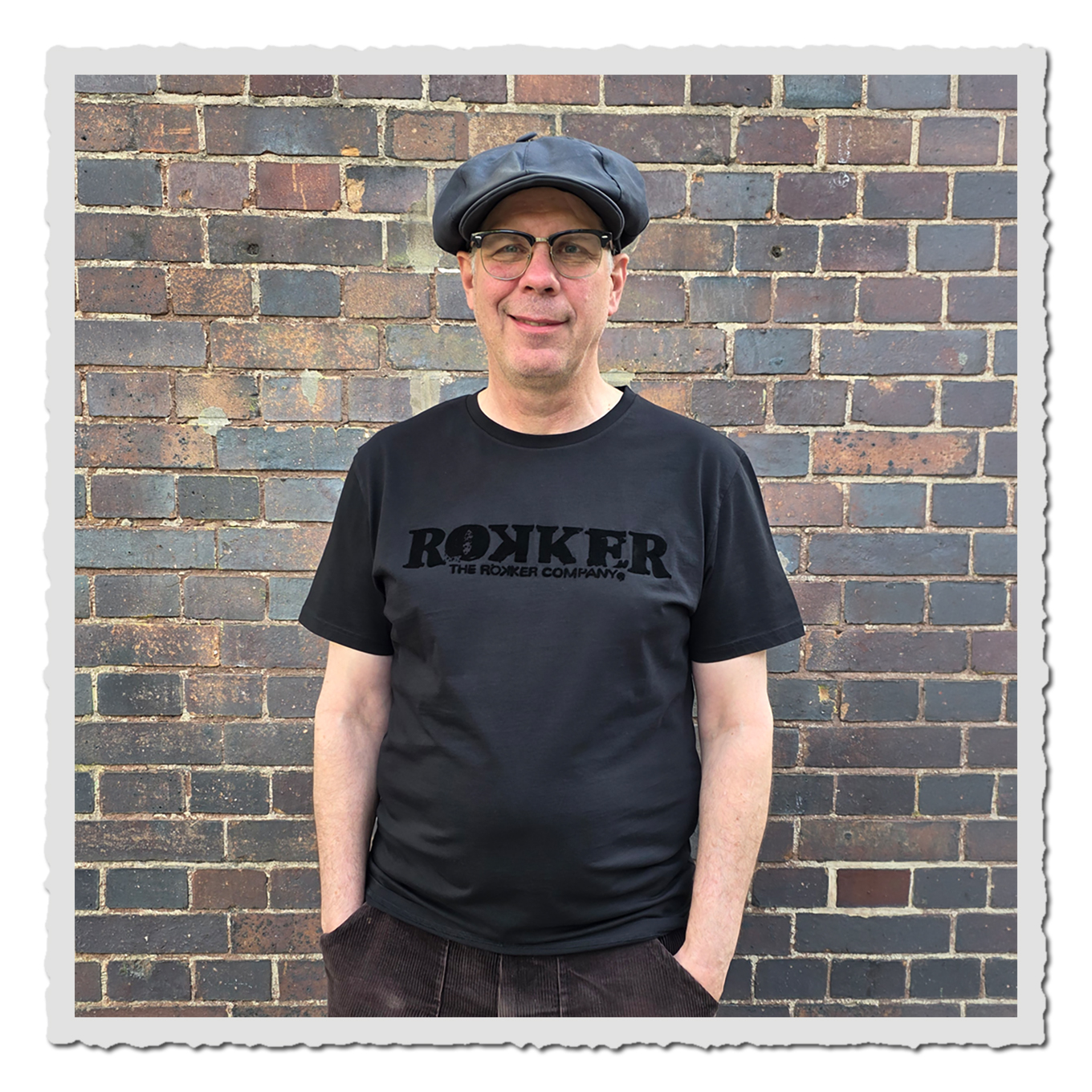 Person trägt schwarzes Rokker T-Shirt und schwarze Kappe vor brauner Backsteinwand, Frontalansicht.